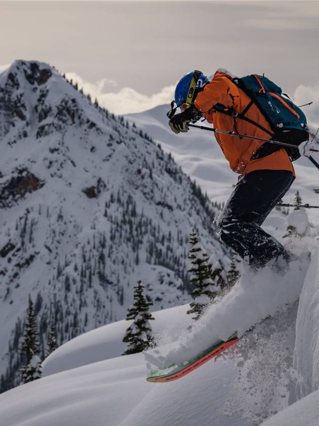 Skier in an orange jacket dropping into deep powder above a snowy mountain ridge