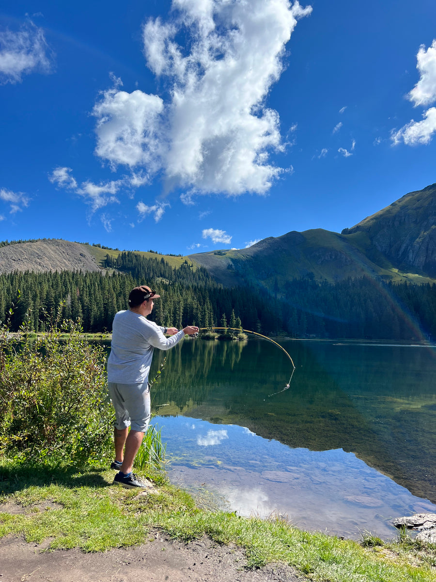 Man casting a fly rod from the grassy shore of a mountain lake under a blue sky