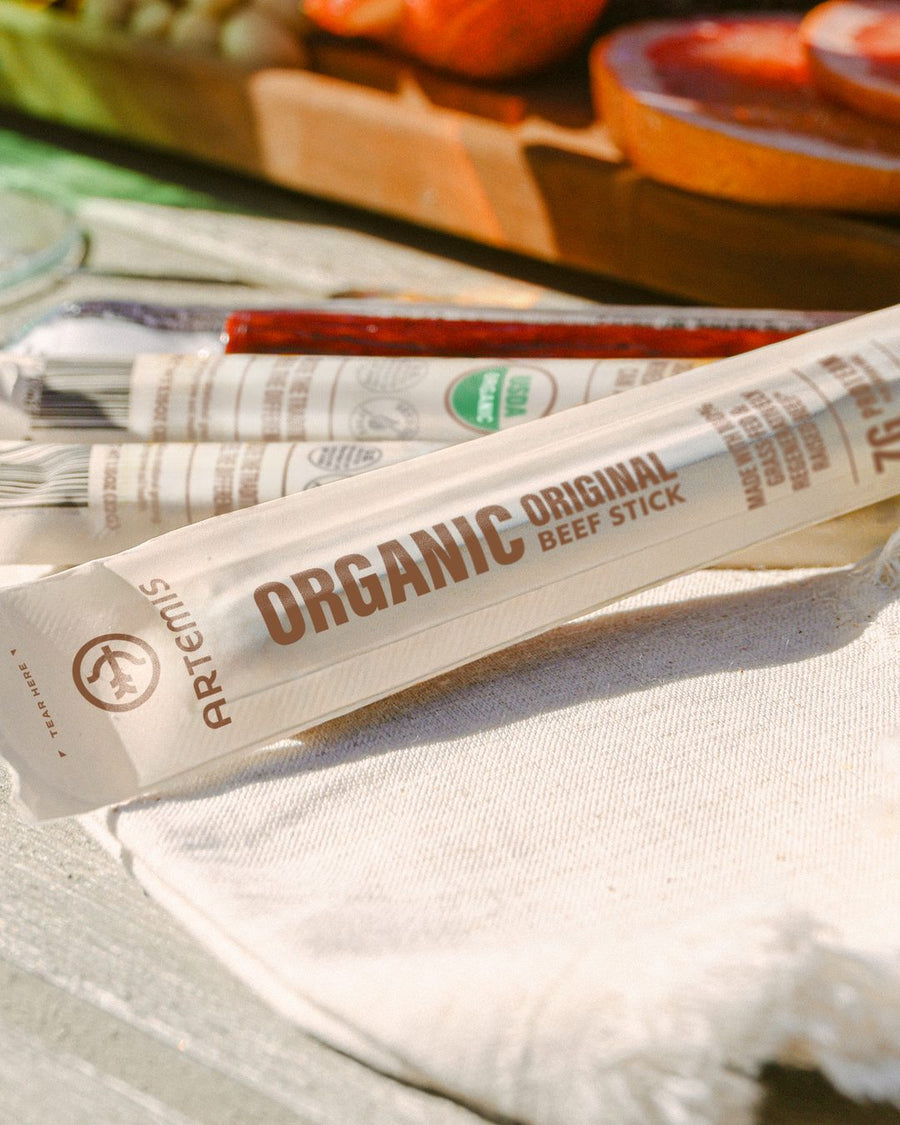 Artemis Organic Original beef sticks on a linen napkin in front of a charcuterie board of fruit on a sunny outdoor table
