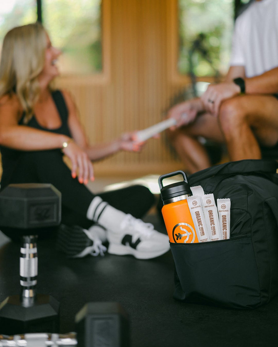 A woman and a man at a home gym passing an Artemis beef stick, a gym bag with an orange bottle and Artemis Organic Original beef sticks on the floor