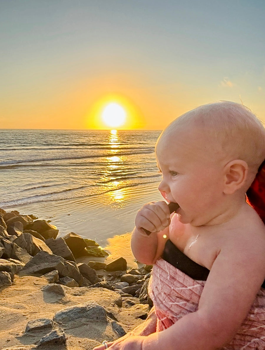 Small child wrapped in a towel on a rocky beach at sunset, chewing an Artemis beef stick