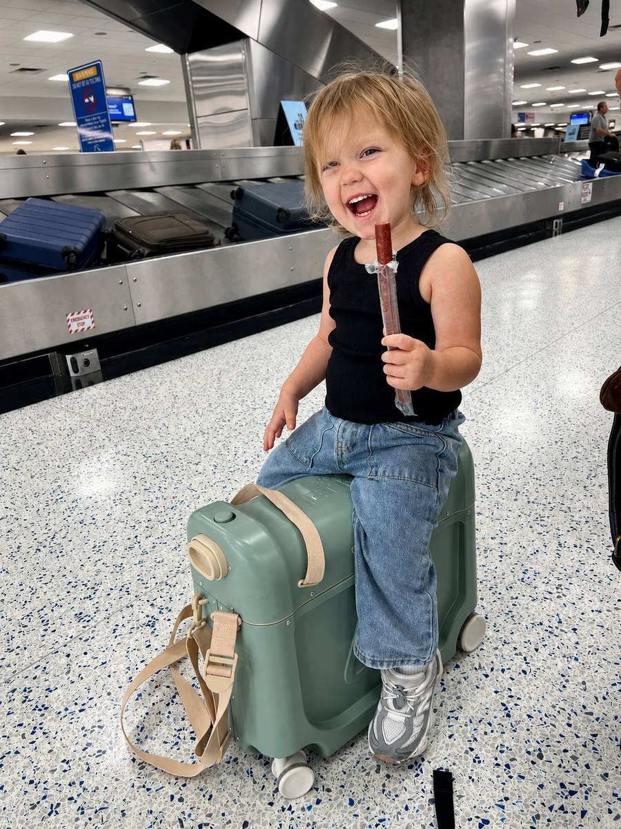 Kid laughing on a ride-on suitcase at an airport baggage claim, holding an Artemis beef stick