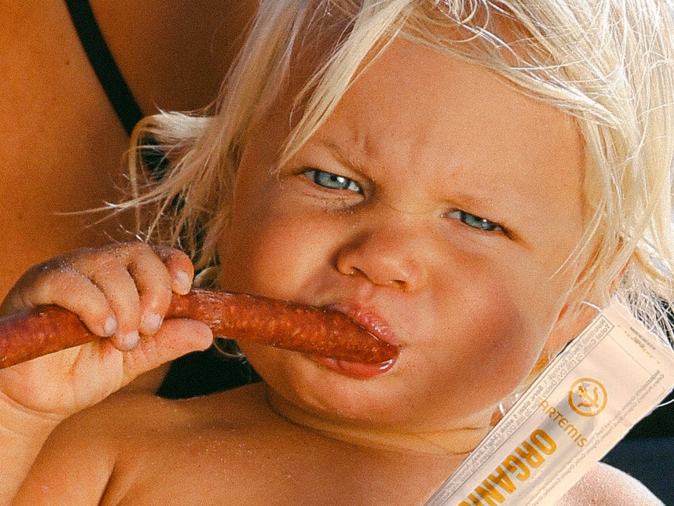 A blonde toddler on a beach bites into an Artemis beef stick while staring at the camera