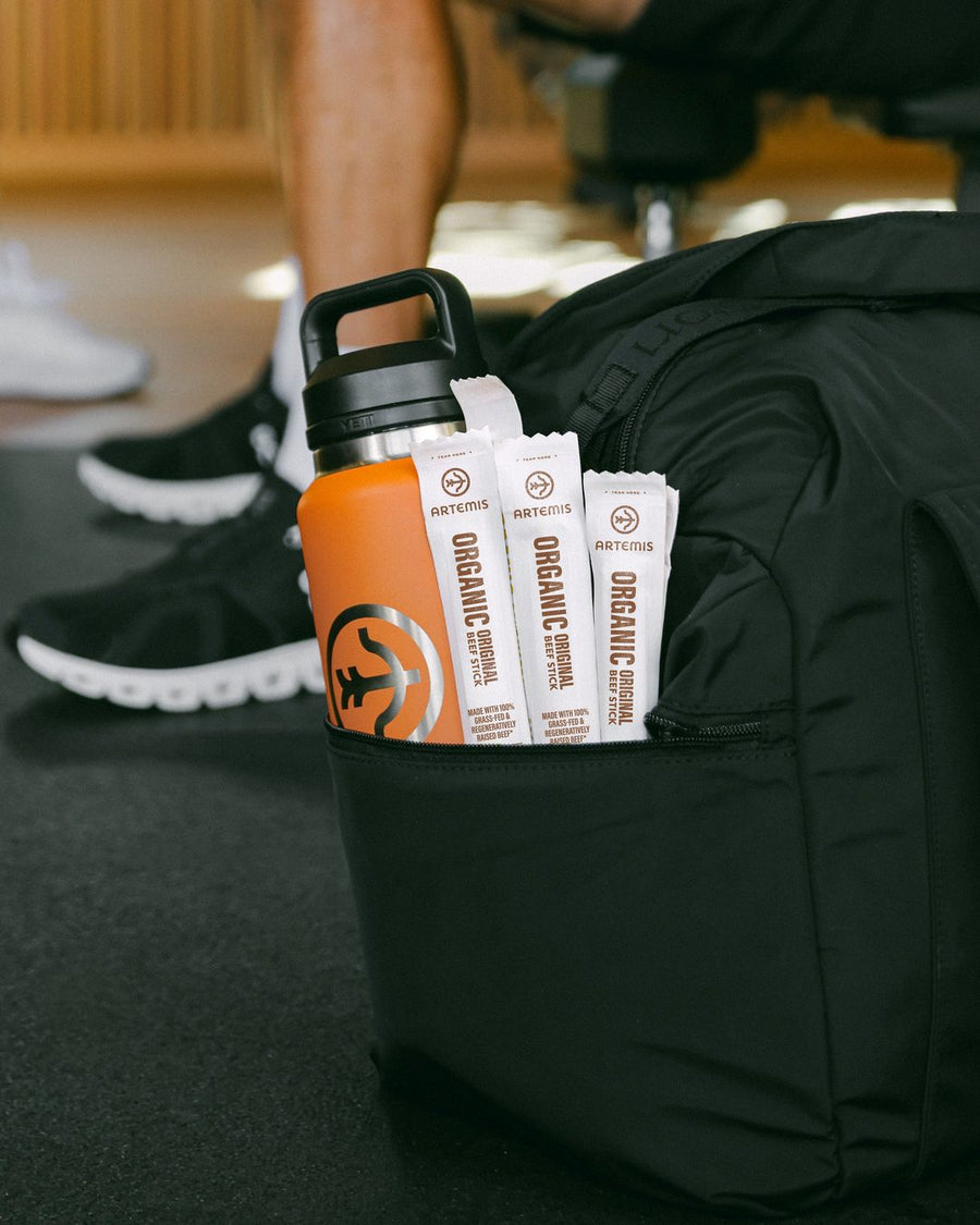 An orange water bottle and three Artemis Organic Original beef sticks tucked in a gym bag on a gym floor, a man on a weight bench behind