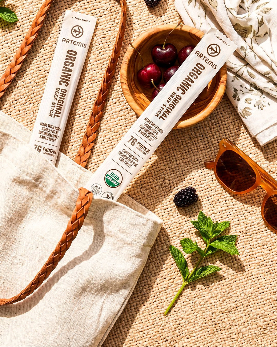 Two Artemis Organic Original beef sticks on a woven picnic blanket beside a linen tote, a bowl of cherries and sunglasses