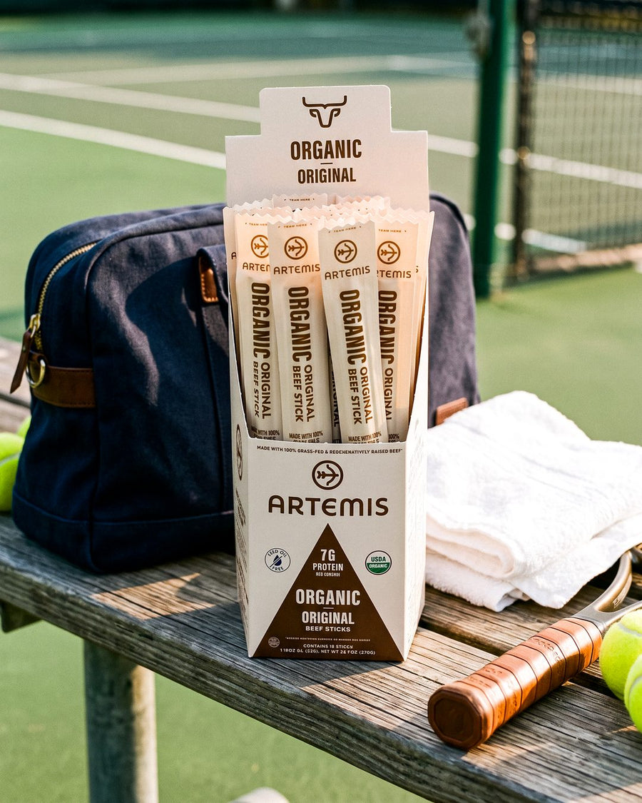 An open box of Artemis Organic Original beef sticks on a courtside bench beside a tennis bag, a towel, a racket and tennis balls